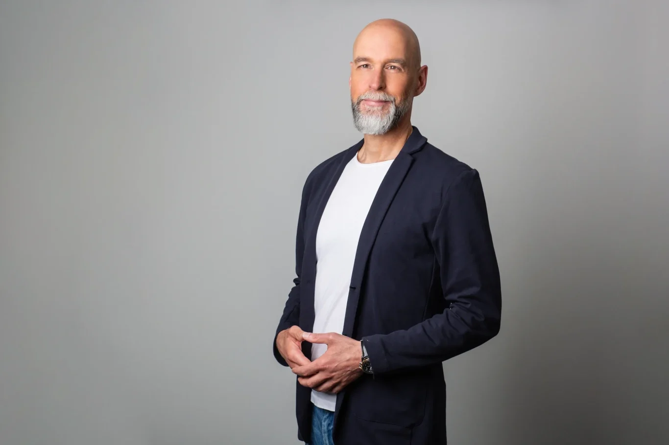 Psychotherapie und Coaching für Klarheit, Selbstwert und Veränderung Mann mit grauem Bart, in einem dunklen Blazer und weißem T-Shirt, steht vor grauem Hintergrund.