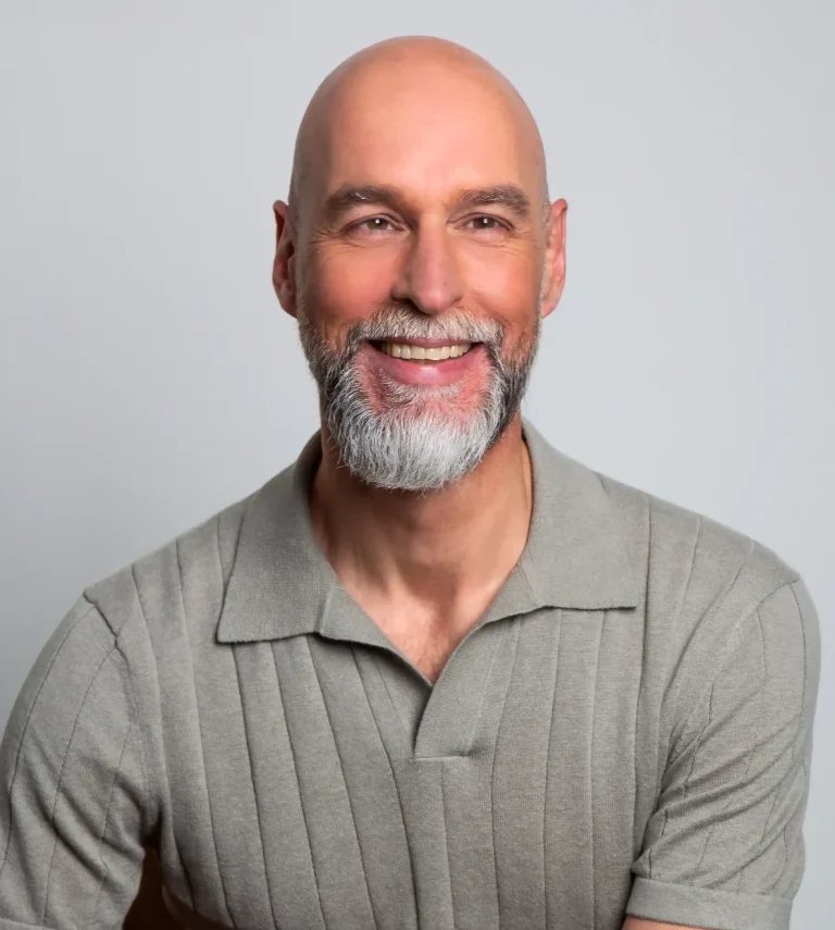 Christian Merlin – Therapeut mit Erfahrung, Präsenz und Klarheit Lächelnder Mann mit grauem Bart und Glatze, trägt ein graues Poloshirt.